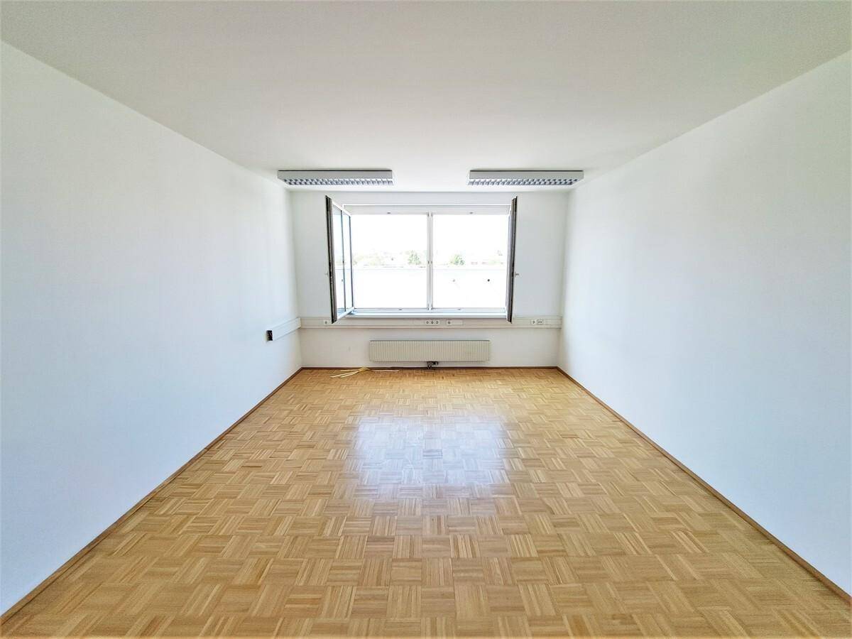 Heller, leerer Raum mit Fischgrätparkettboden und einem großen Fenster, das viel Tageslicht hereinlässt.