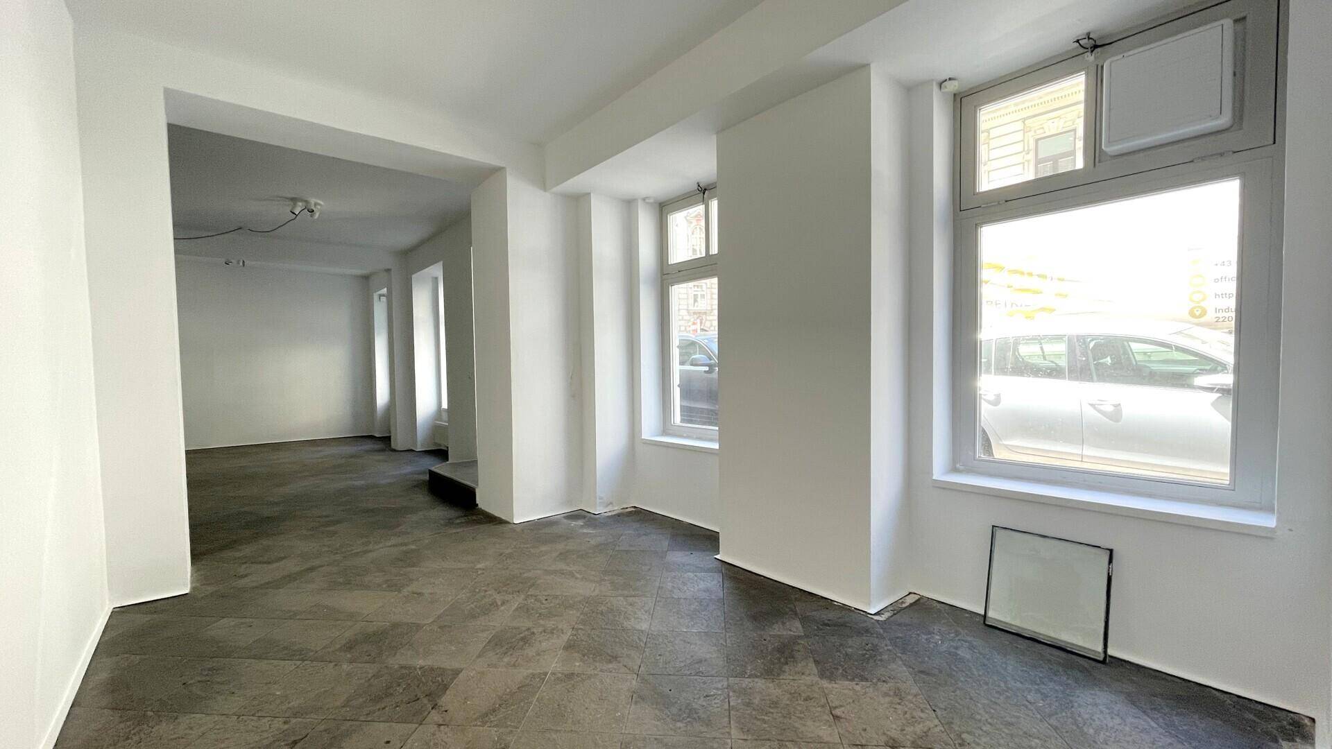 Geräumiger, heller Raum mit großen Fenstern zur Straße und grauen Fliesen, ideal für ein Geschäft oder Büro.