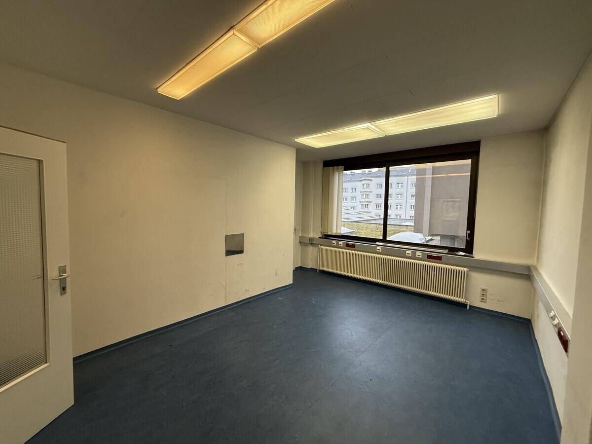 Heller Büroraum mit blauen Vinylböden, großem Fenster und Blick auf umliegende Gebäude.