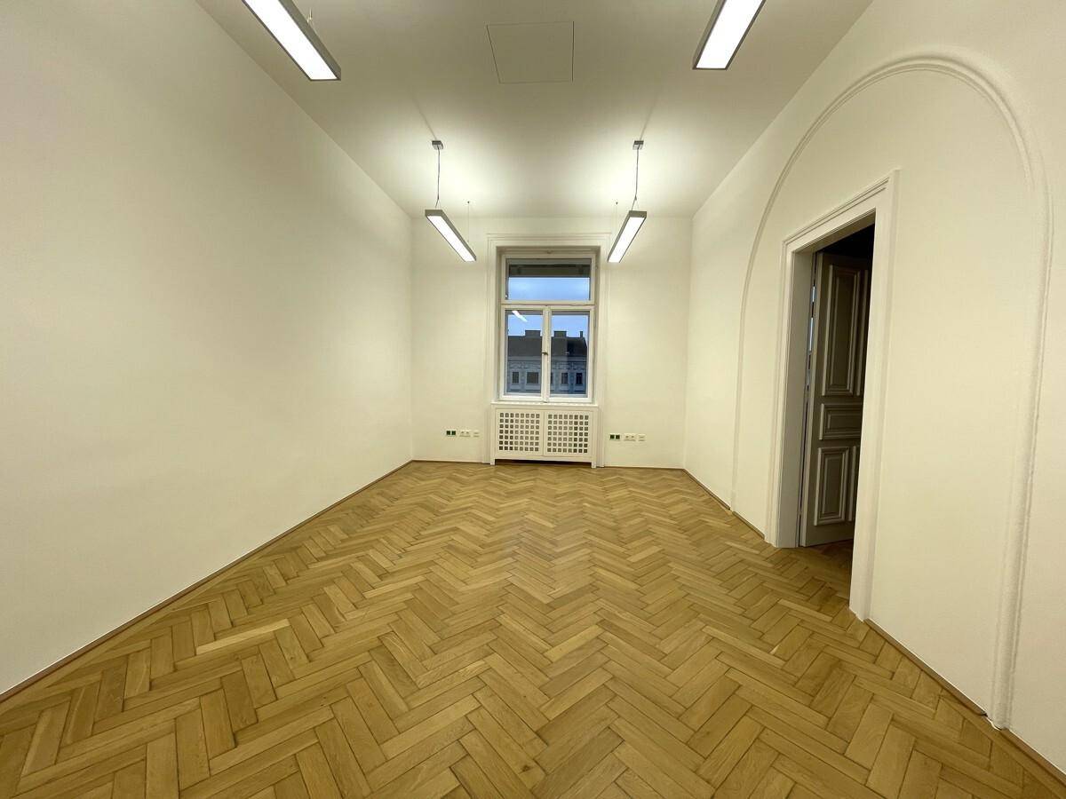 Geräumiger Büroraum mit Fischgrätparkett, einem großen Fenster und einem eleganten Türbogen.