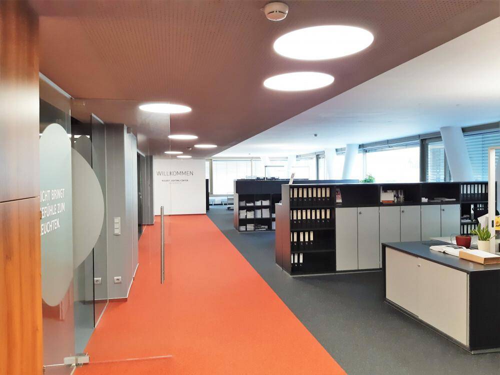 Heller Büroflur mit orangefarbenem Teppich und runden Deckenleuchten, führt zu offenen Arbeitsbereichen mit Aktenschränken.