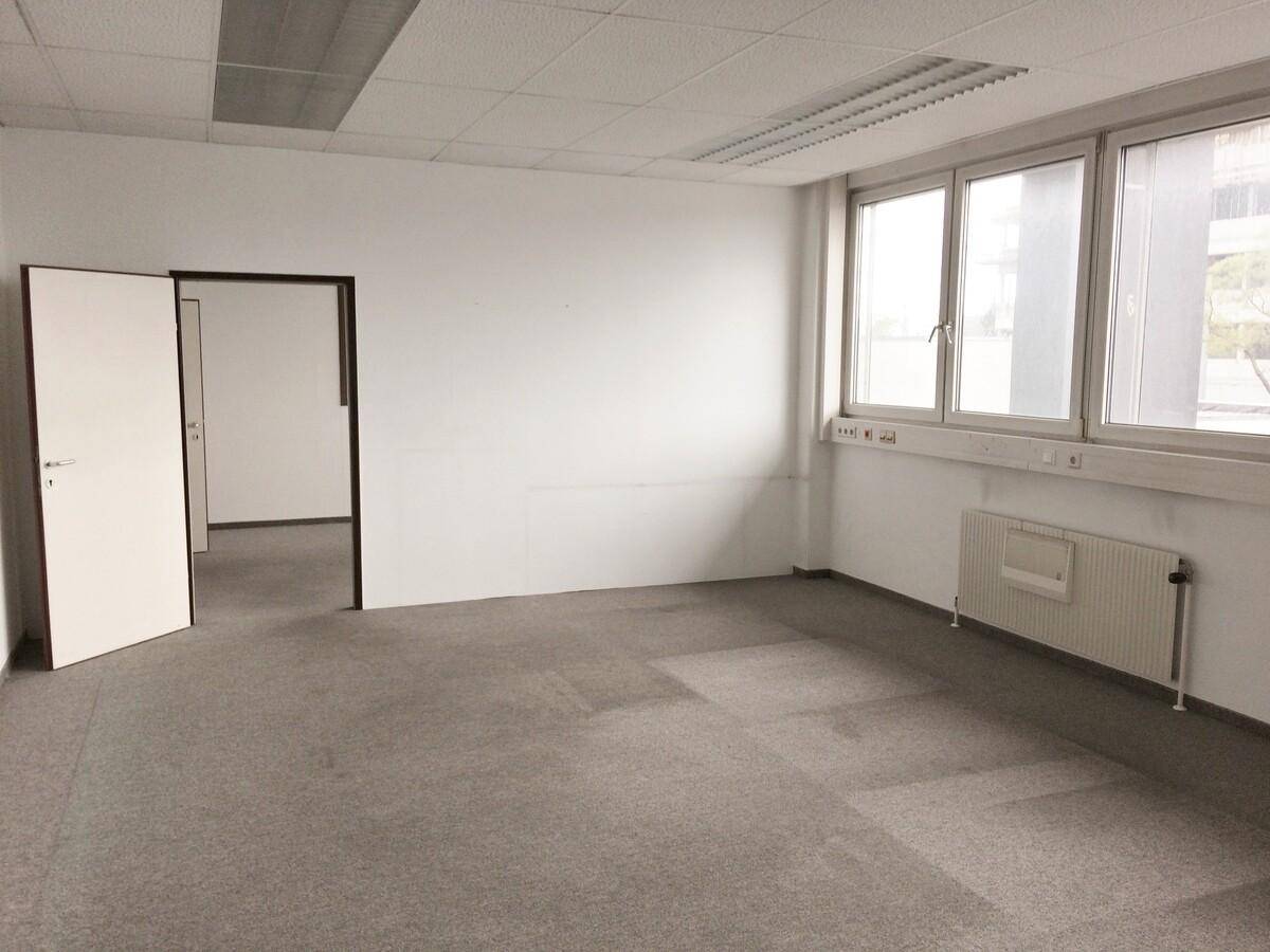 Leeres Büro mit grauem Teppichboden, weißen Wänden und großen Fenstern, die Tageslicht hereinlassen.
