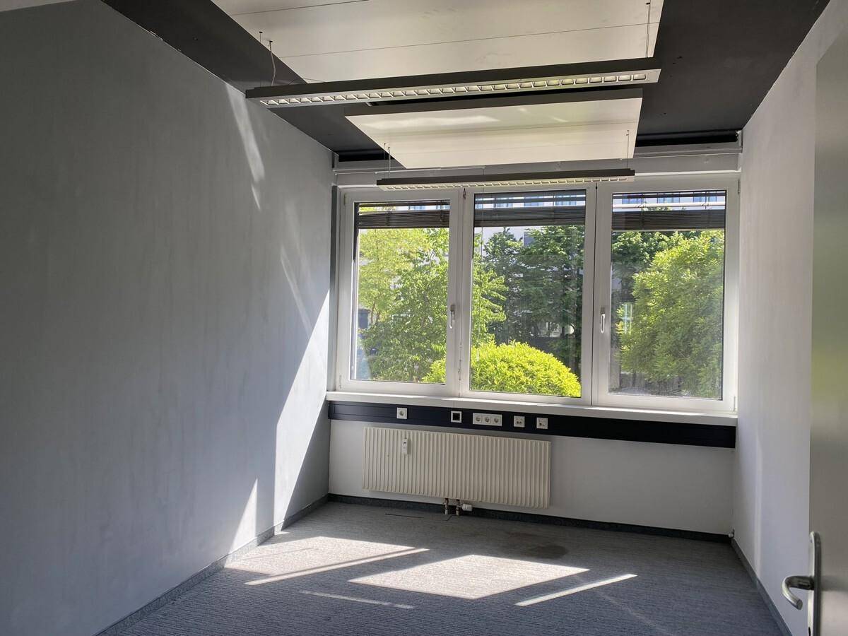 Helles Einzelbüro mit Teppichboden, großen Fenstern und Blick auf grüne Bäume.