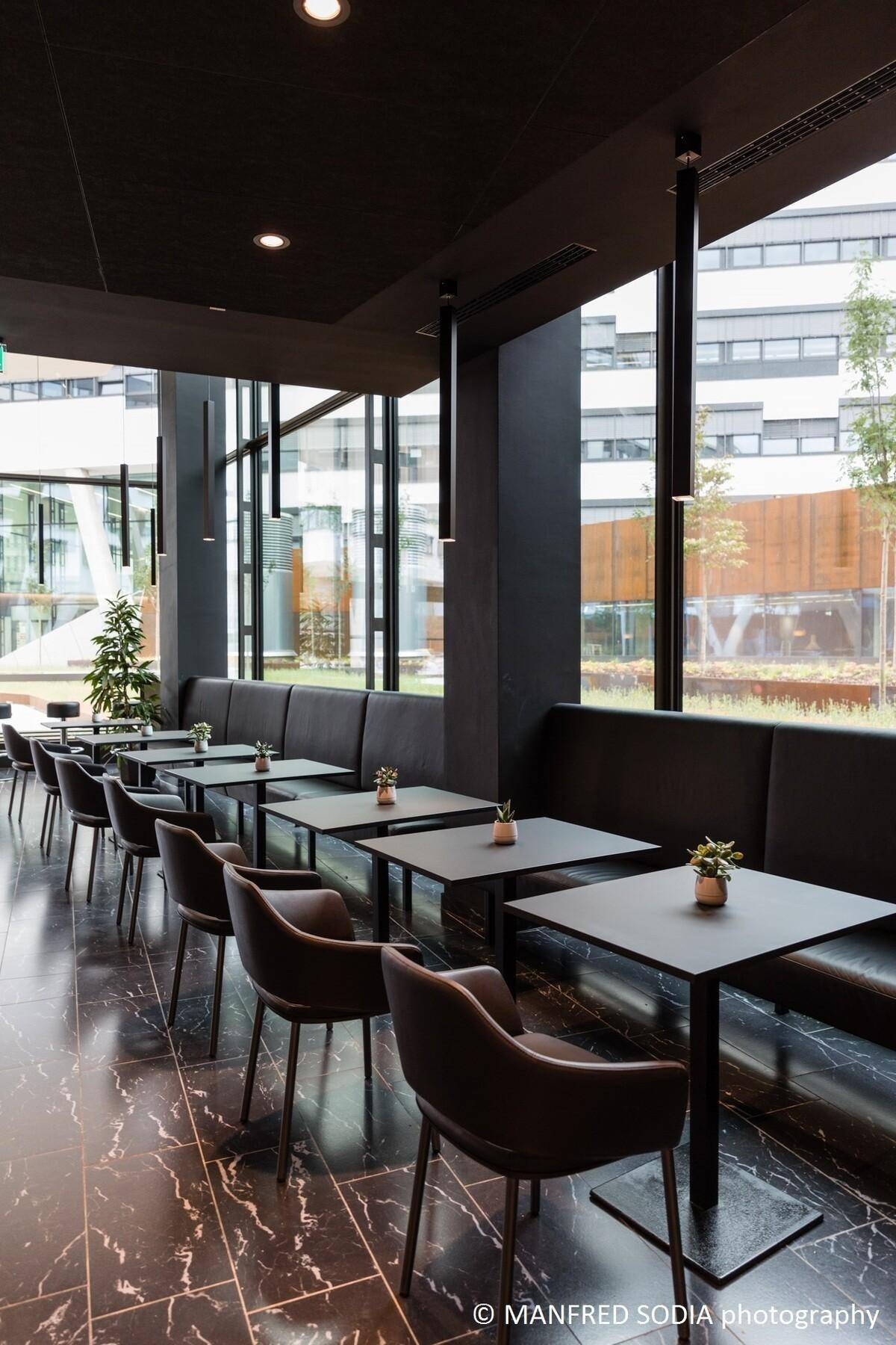 Modernes Café mit dunklen Tischen, bequemen Sitzbänken und großen Fenstern mit Blick auf den Außenbereich.