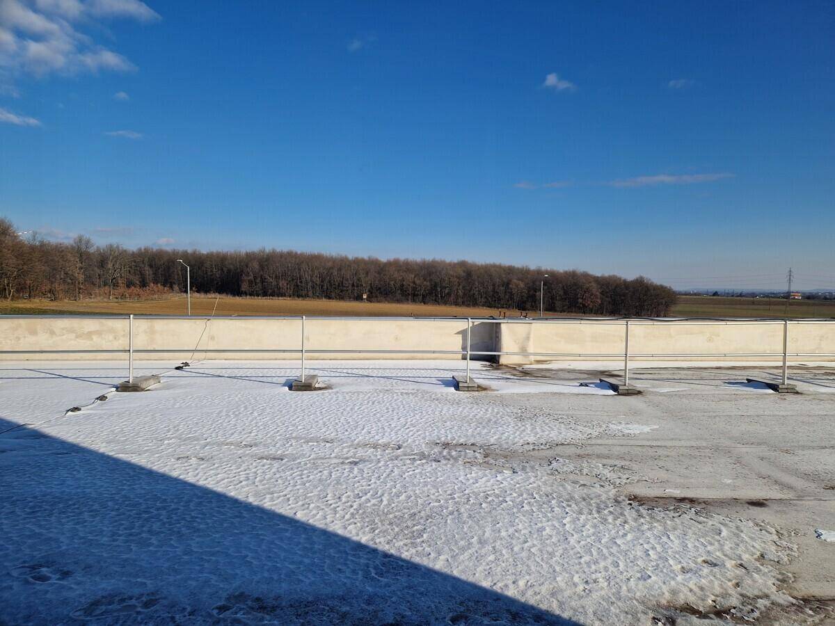 Flachdach mit Schneebedeckung, einer niedrigen Mauer und einer Baumreihe am Horizont unter blauem Himmel.
