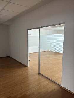 Heller Büroraum mit Holzboden und einer modernen Glaswand, die einen weiteren Raum abtrennt.