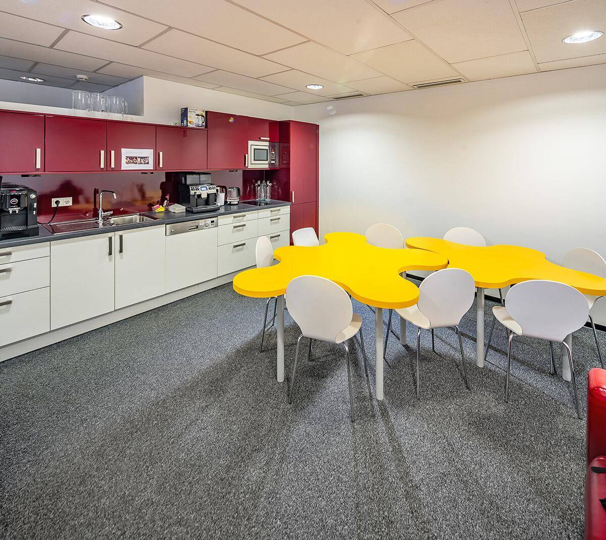 Moderne Küche mit roten Schränken, weißen Arbeitsplatten und einem auffälligen gelben Tisch, ideal für gemeinsame Pausen.