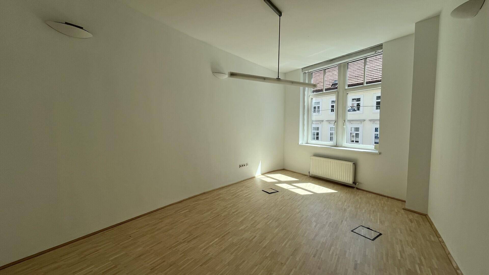 Heller Büroraum mit Holzfußboden, einem Fenster mit Jalousien und einer Deckenleuchte für gute Beleuchtung.