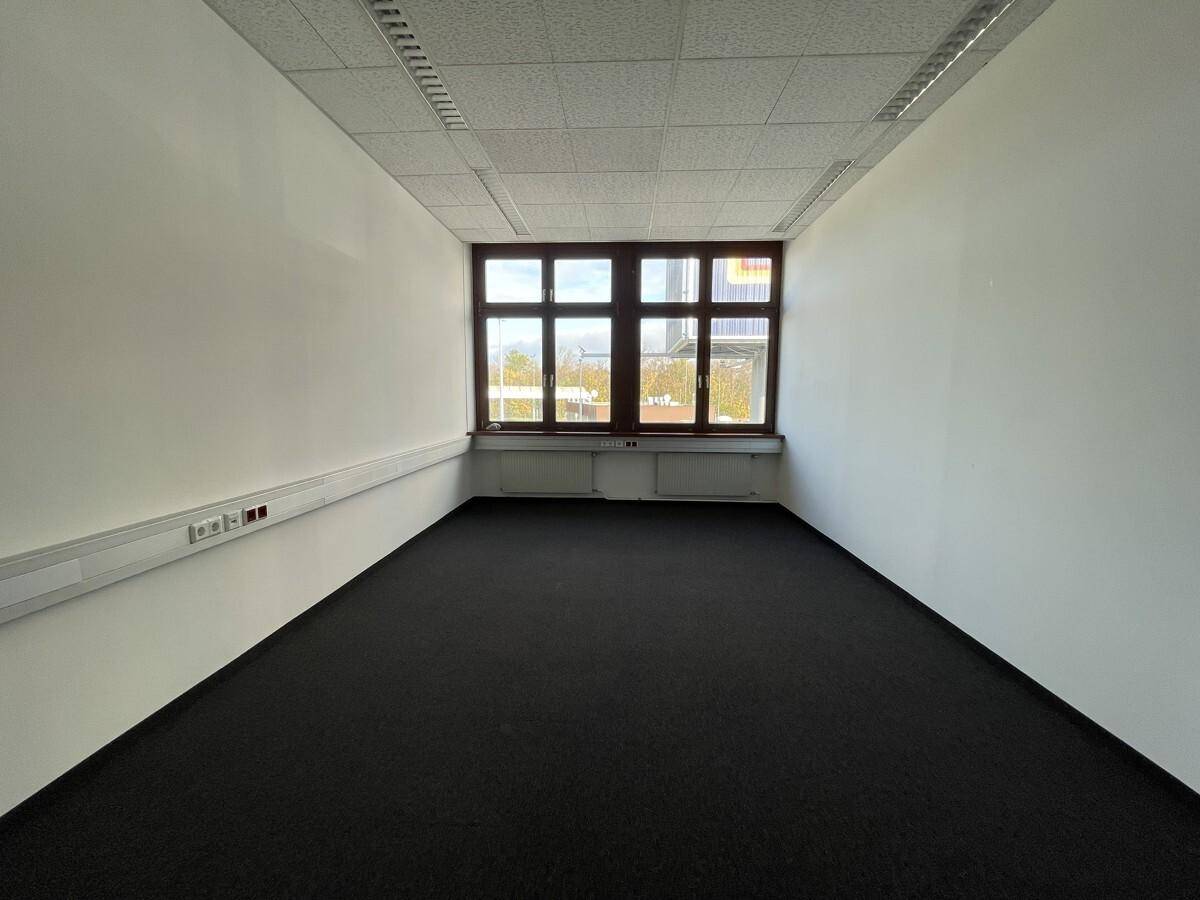 Schmaler Büroraum mit Teppichboden, weißen Wänden und einem großen Fenster für ausreichend Tageslicht.