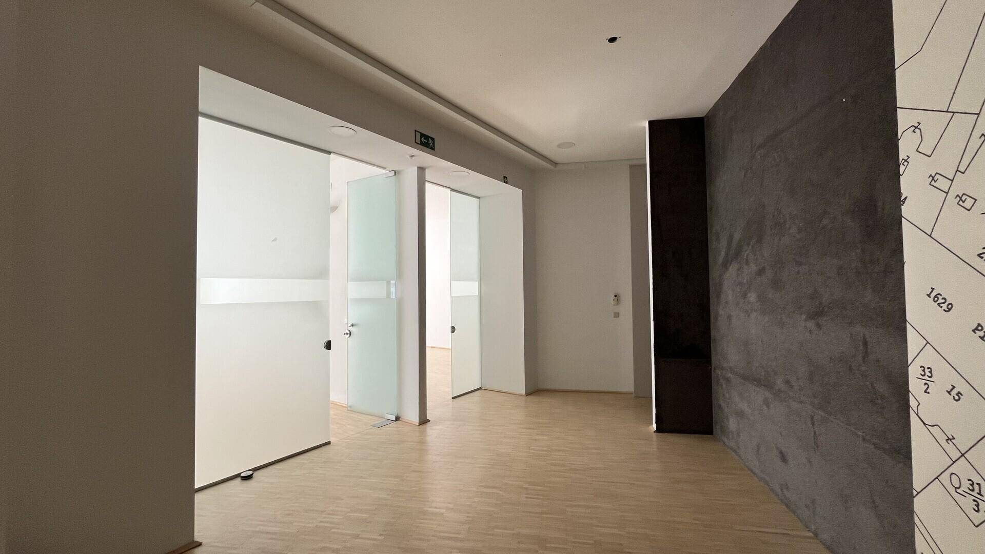 Heller Flur oder Bürobereich mit Holzfußboden, modernen Glasschiebetüren und einer dunklen Akzentwand.