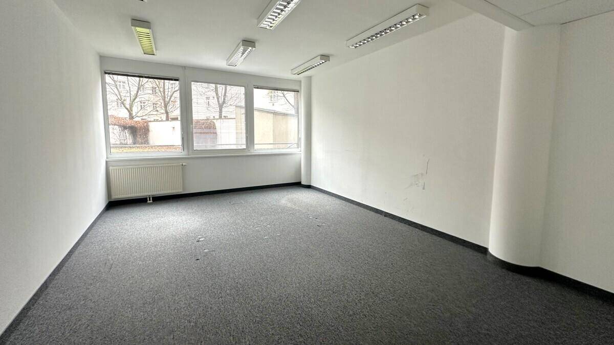 Geräumiger, leerer Büroraum mit Teppichboden und mehreren Fenstern, die eine helle Arbeitsumgebung schaffen.