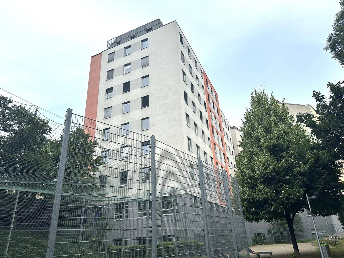 Hohes Wohngebäude mit moderner Fassade, vielen Fenstern und einem Zaun im Vordergrund.