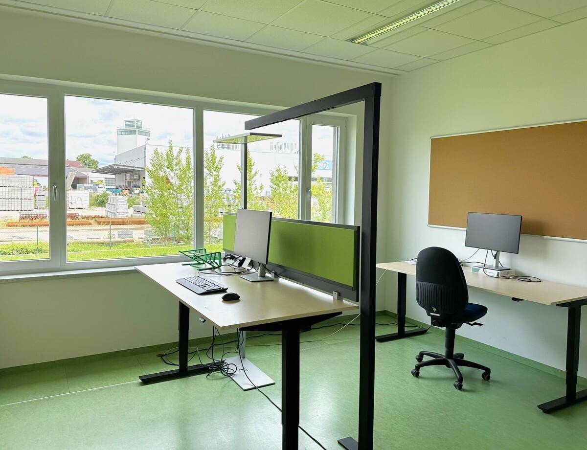 Helles Büro mit zwei modernen Steh-Sitz-Schreibtischen und großen Fenstern mit Blick ins Grüne.