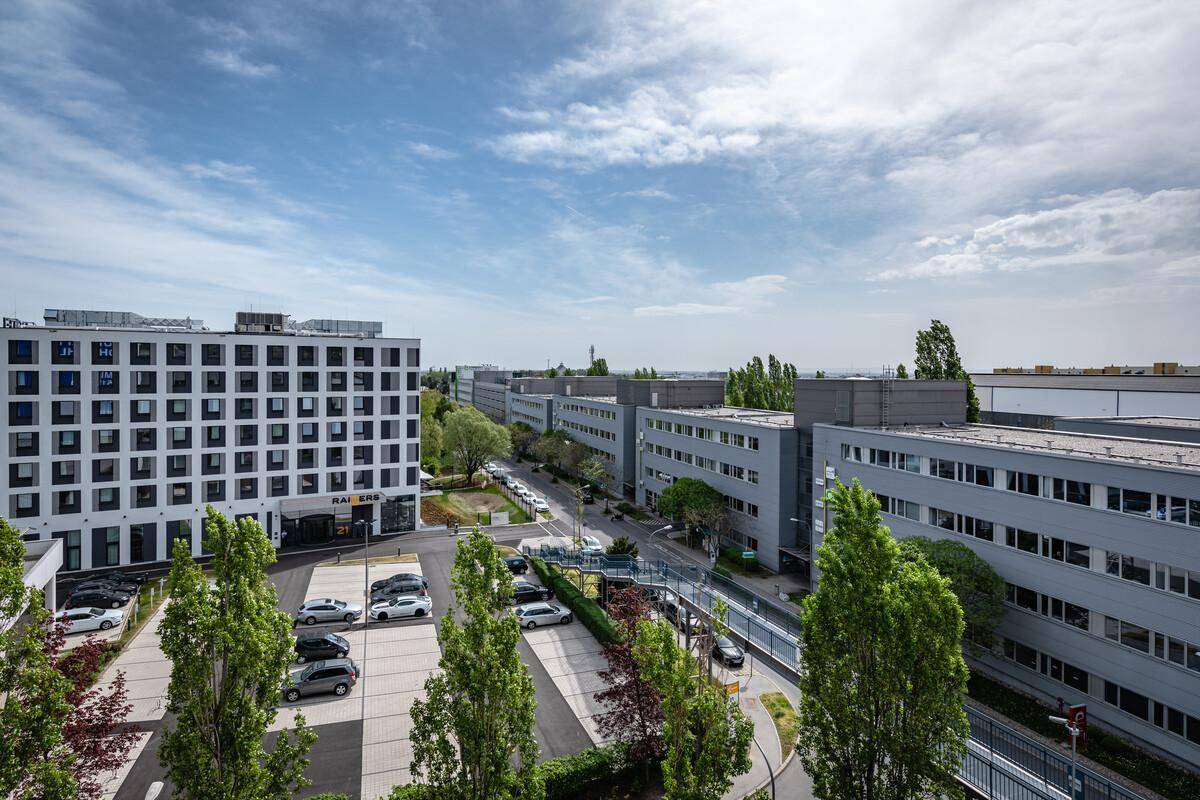 Panoramablick auf einen modernen Bürokomplex mit Parkplätzen und grünen Bäumen unter einem teilweise bewölkten Himmel.