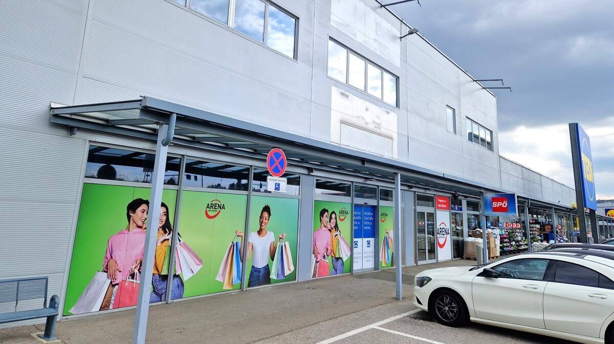 Eingangsbereich eines Einkaufszentrums mit großen Schaufenstern und Werbeplakaten, davor ein Parkplatz.