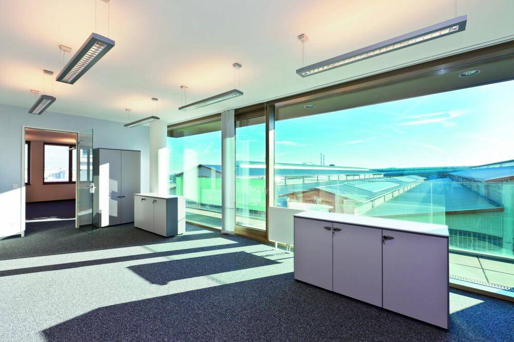 Geräumiges, leeres Büro mit großen Fenstern, die viel Tageslicht und einen weiten Ausblick auf Industriegebäude bieten.