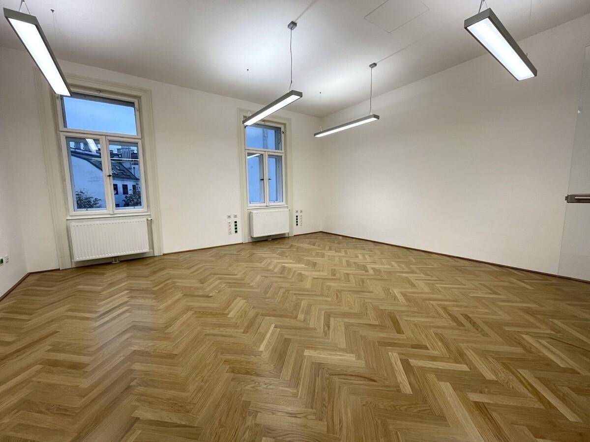 Heller Büroraum mit Fischgrätparkett und zwei großen Fenstern, die den Raum mit Tageslicht füllen.