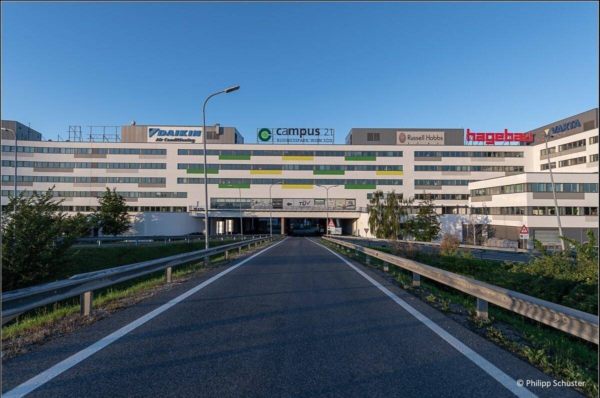 Weitläufige Ansicht eines großen Bürogebäudes mit verschiedenen Firmenlogos und einer Zufahrtsstraße im Vordergrund.