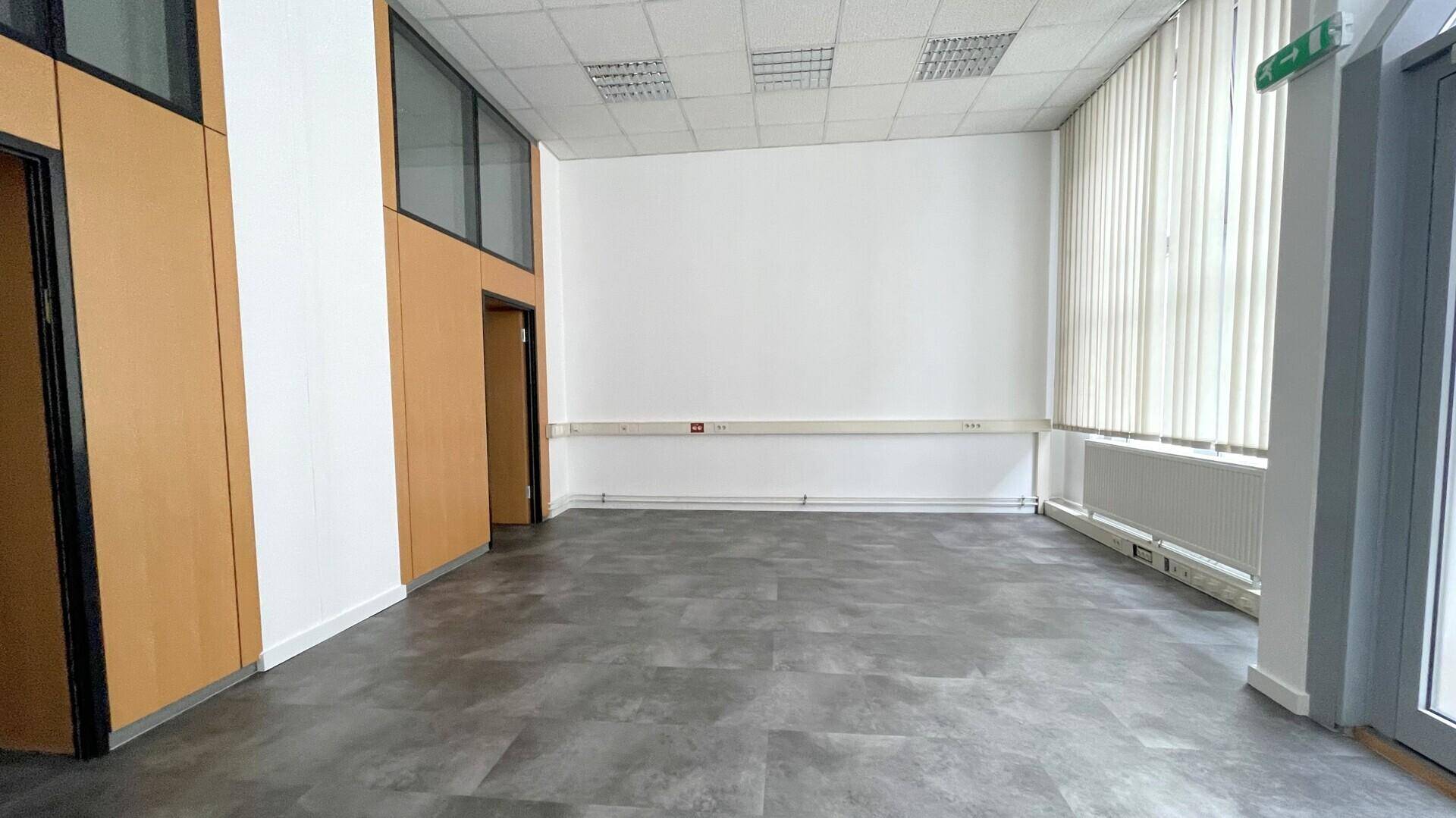 Geräumiger Büroraum mit dunklem Vinylboden, weißen Wänden und Holzvertäfelungen an den Türen und Fenstern.