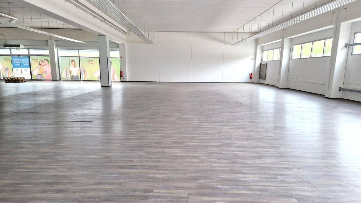 Großer, leerer Gewerberaum mit hellem Holzboden, weißen Wänden und sichtbaren Deckeninstallationen.