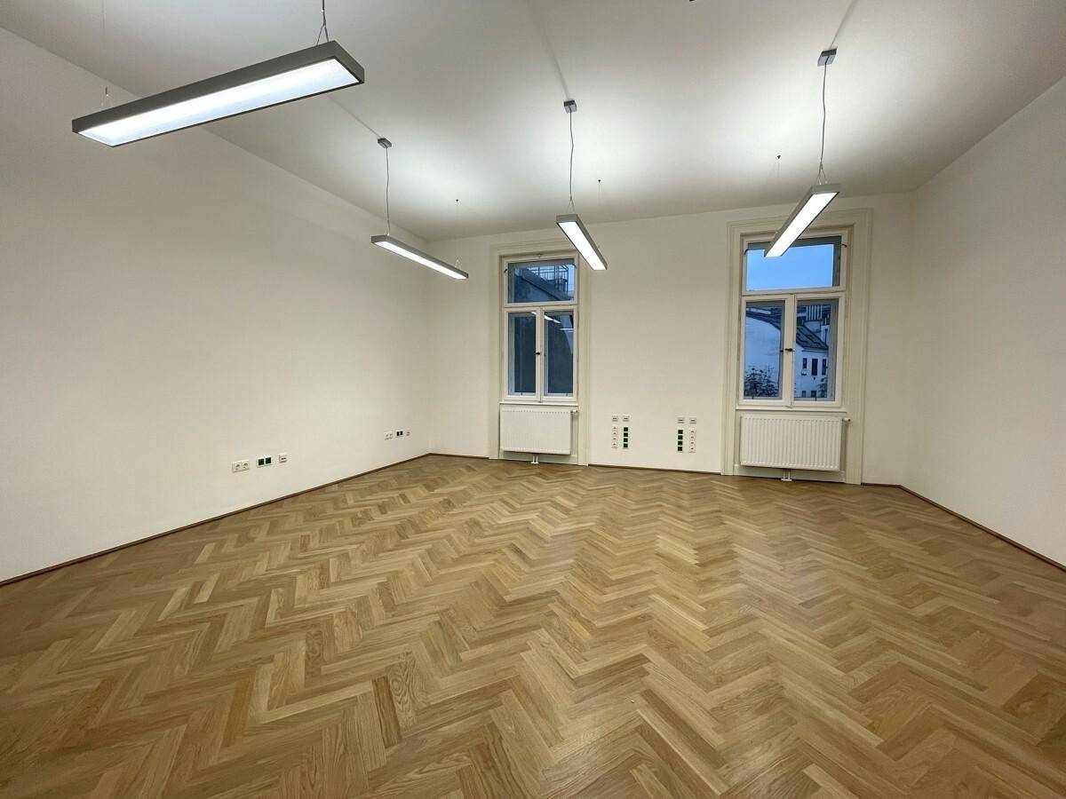 Leerer Büroraum mit Fischgrätparkett und zwei großen Fenstern, die für eine angenehme Helligkeit sorgen.