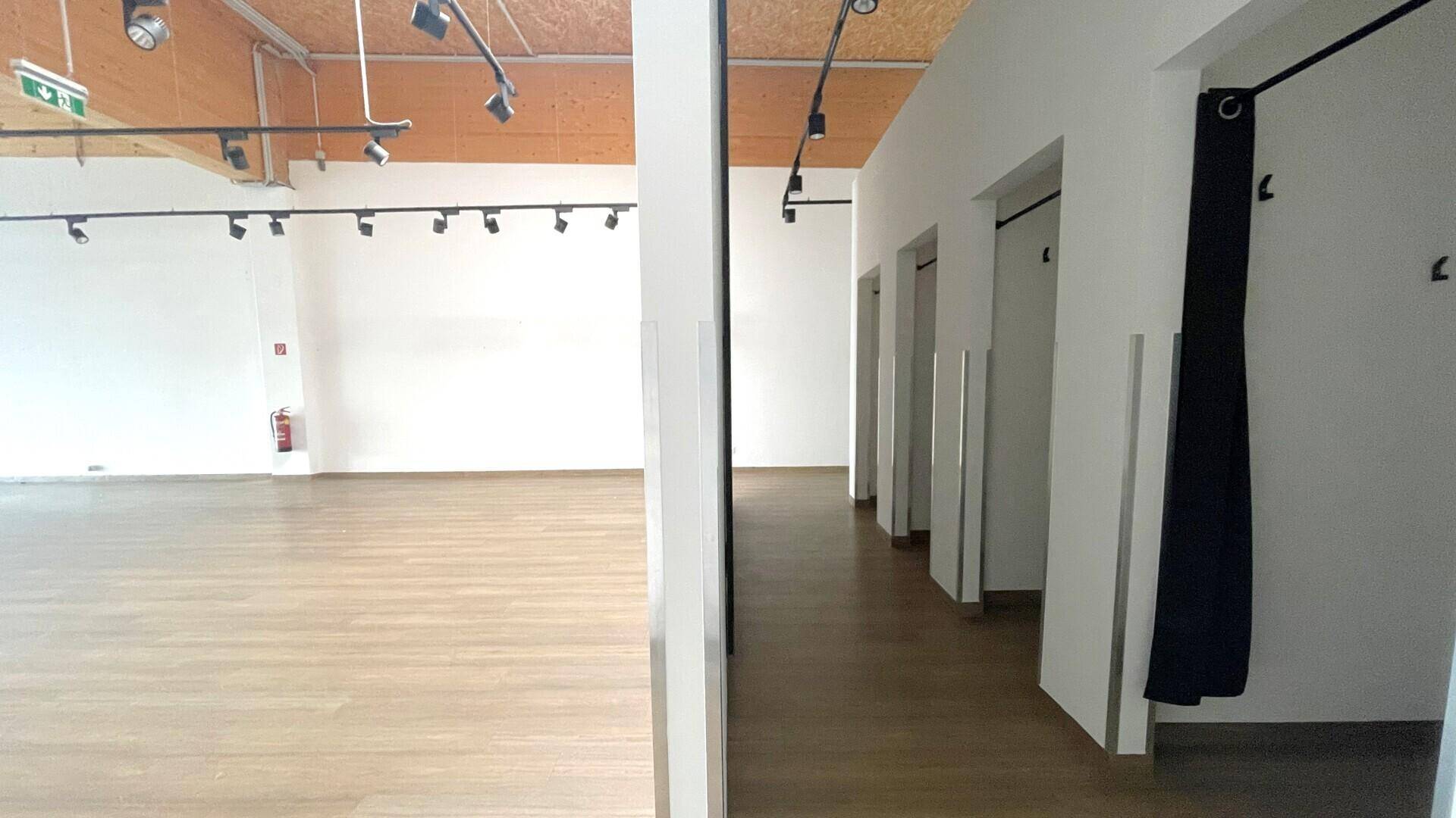 Heller Verkaufsraum mit Holzbalkendecke, Schienenbeleuchtung und einer Reihe von Umkleidekabinen mit Vorhängen.