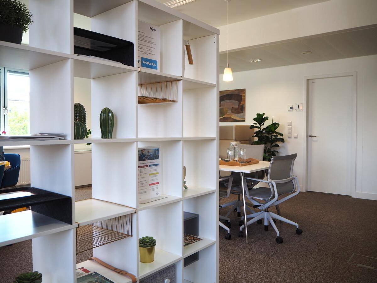 Offener Bürobereich mit Raumteiler, Regalen und Blick auf einen Konferenzbereich, fördert flexible Arbeitsweisen.