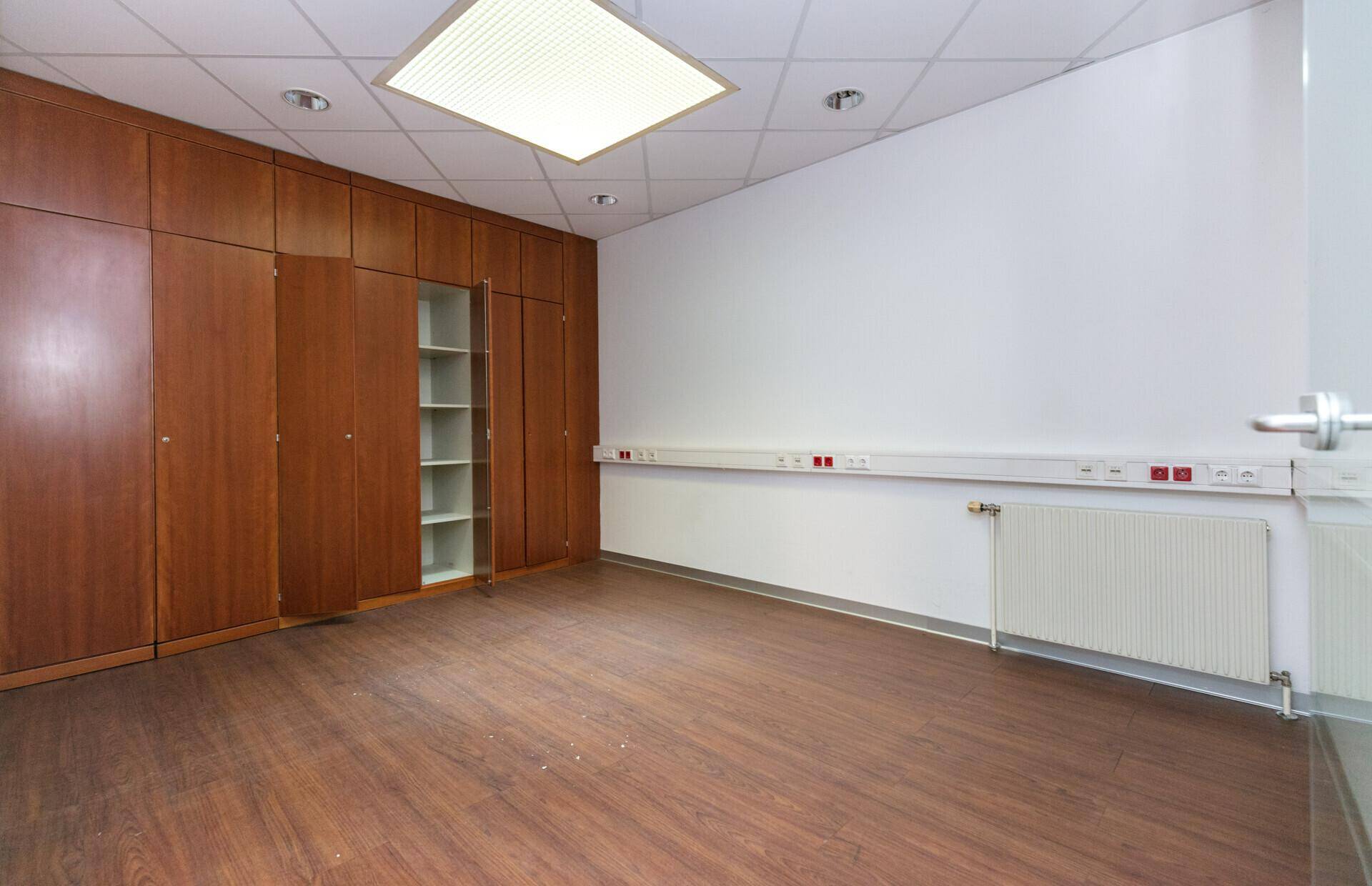 Büroraum mit dunklen Holzschränken entlang einer Wand, Laminatboden und Heizkörper.