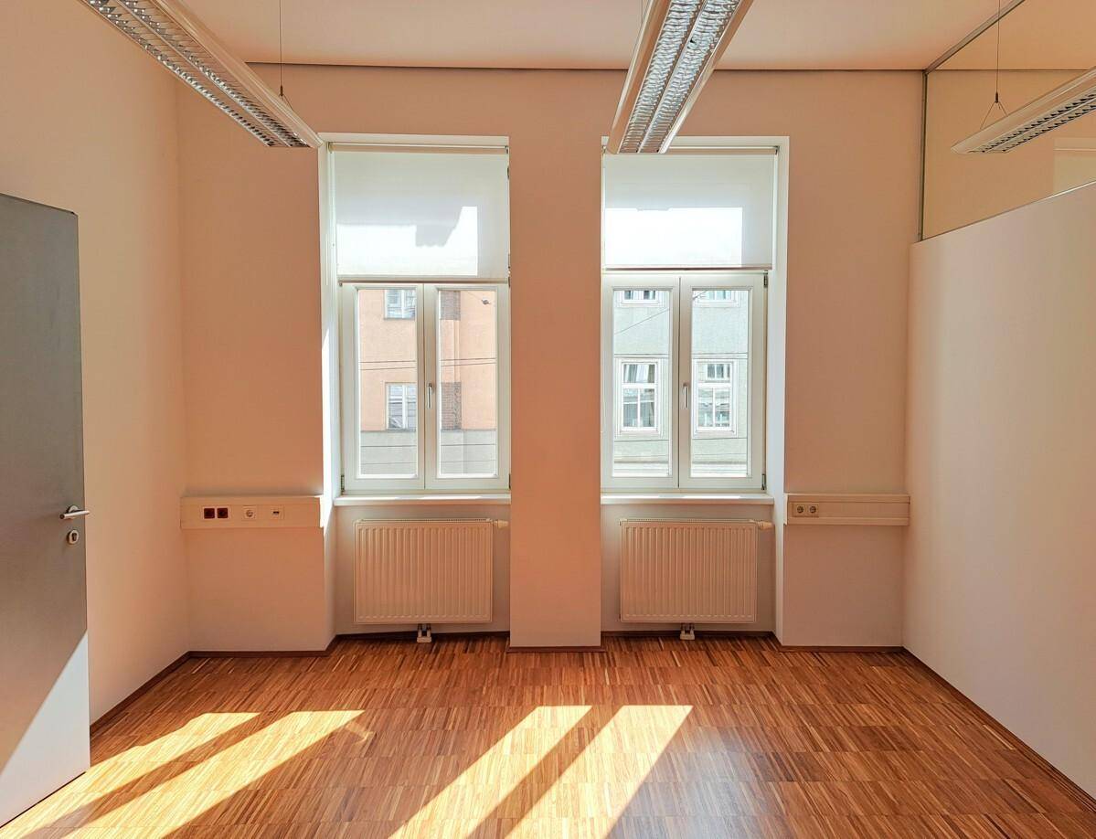 Leeres Büro mit zwei hohen Fenstern, Heizkörpern und einem warmen Holzfußboden, sehr hell.