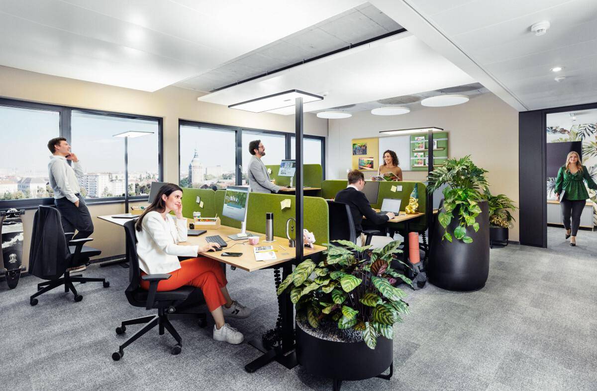 Helles Großraumbüro mit ergonomischen Arbeitsplätzen, grünen Trennwänden und Blick auf die Stadt.