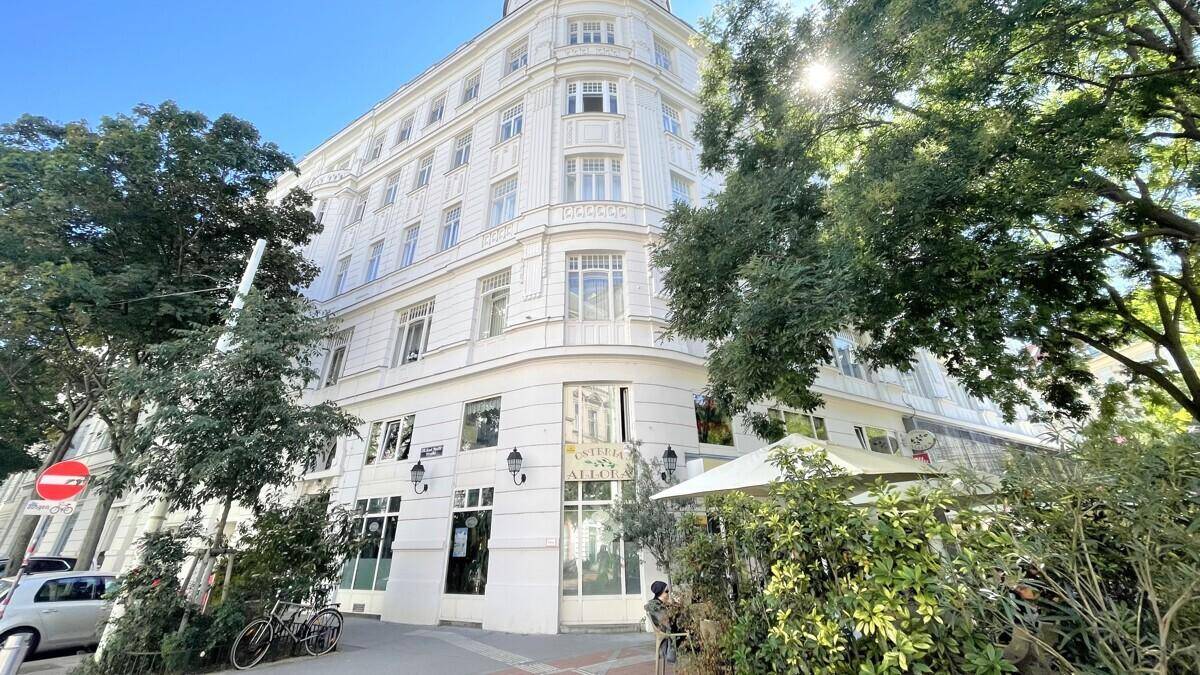 Historisches Eckgebäude mit Geschäften im Erdgeschoss und Bäumen davor an einem sonnigen Tag.