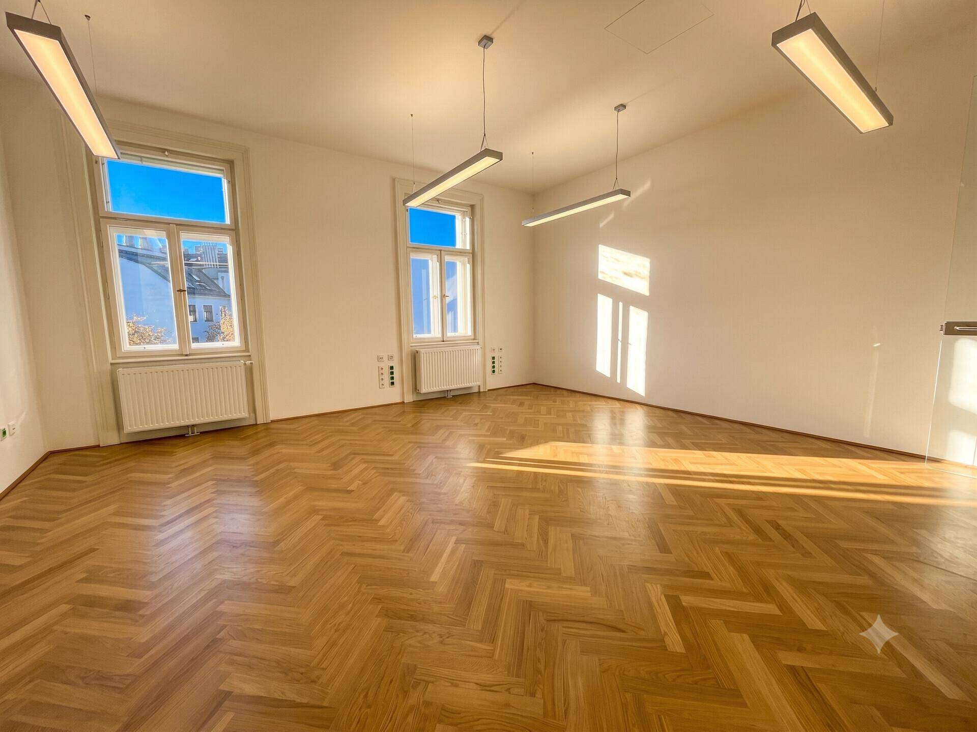 Großer, leerer Büroraum mit elegantem Fischgrätparkett und hohen Fenstern, die den Raum erhellen.