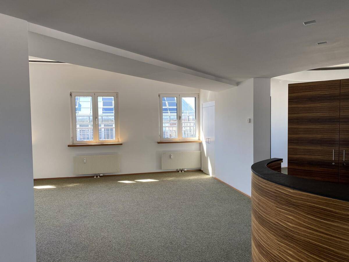Geräumiger Büroraum mit Teppichboden, zwei Fenstern und einer modernen Holzvertäfelung an der Seite.