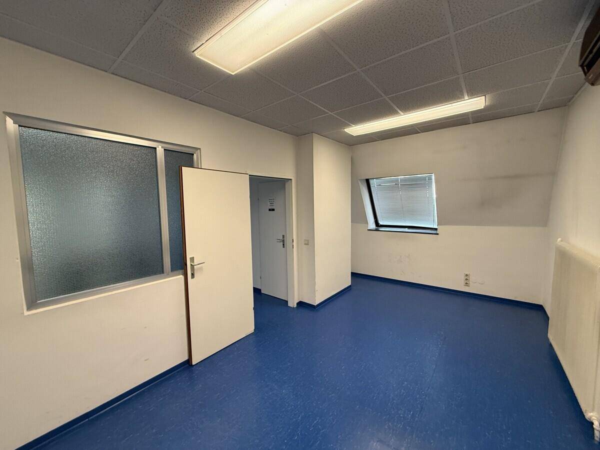 Kleiner Büroraum mit blauen Vinylböden, einem Fenster und Zugang zu einem weiteren Bereich.