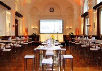 Großer Konferenzsaal mit Holzboden, Projektionsfläche und modernen Stehtischen, ideal für Veranstaltungen und Präsentationen.