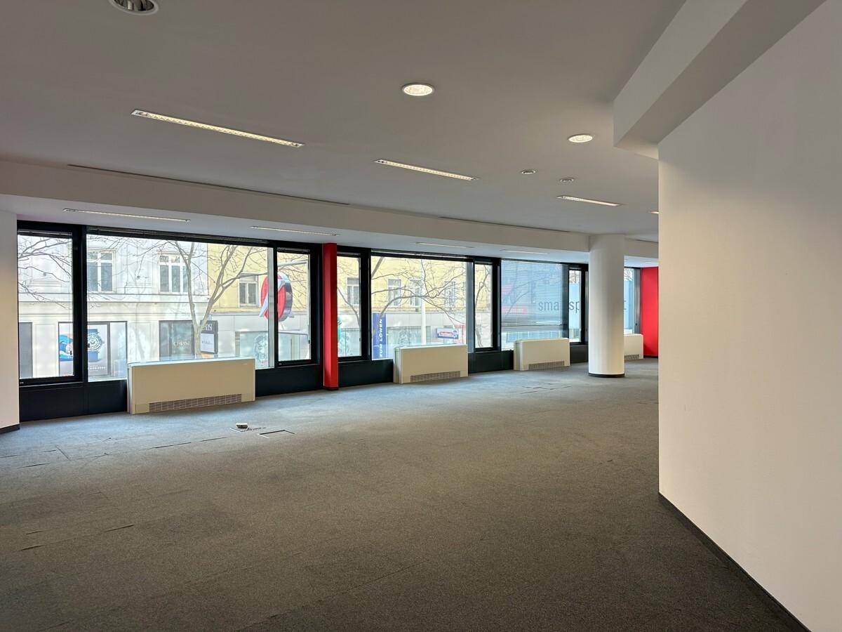Heller Büroraum mit großen Fenstern, die einen Blick auf die Straße bieten, und grauem Teppichboden.