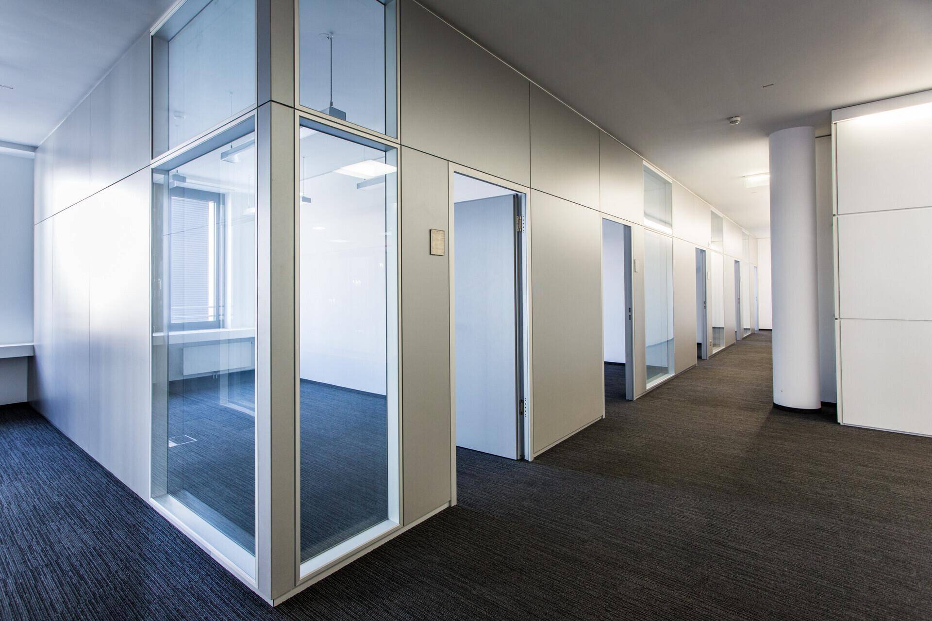 Langer Bürogang mit Teppichboden, Glasbüros und weißen Wänden, der eine offene Atmosphäre schafft.