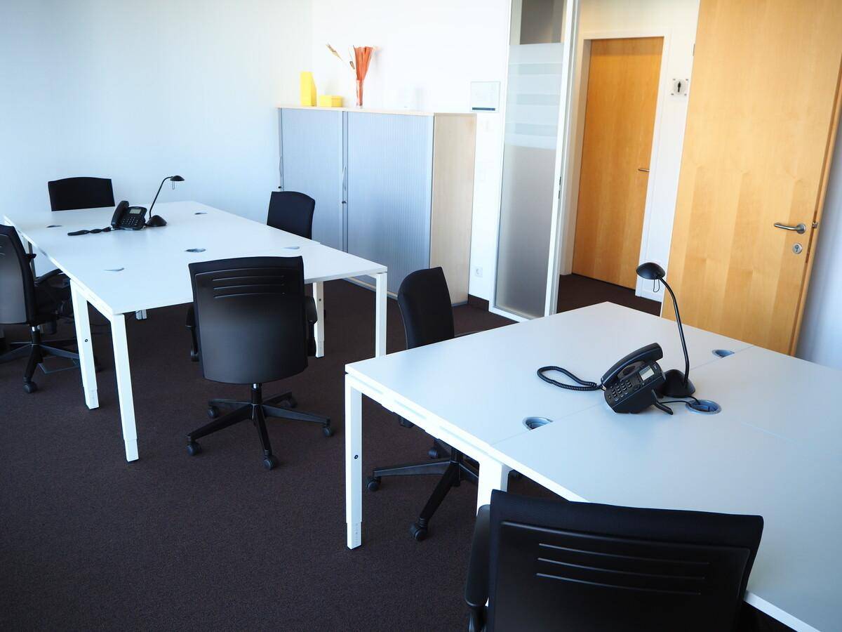 Großraumbüro mit mehreren Arbeitsplätzen, weißen Schreibtischen und dunklem Teppichboden.