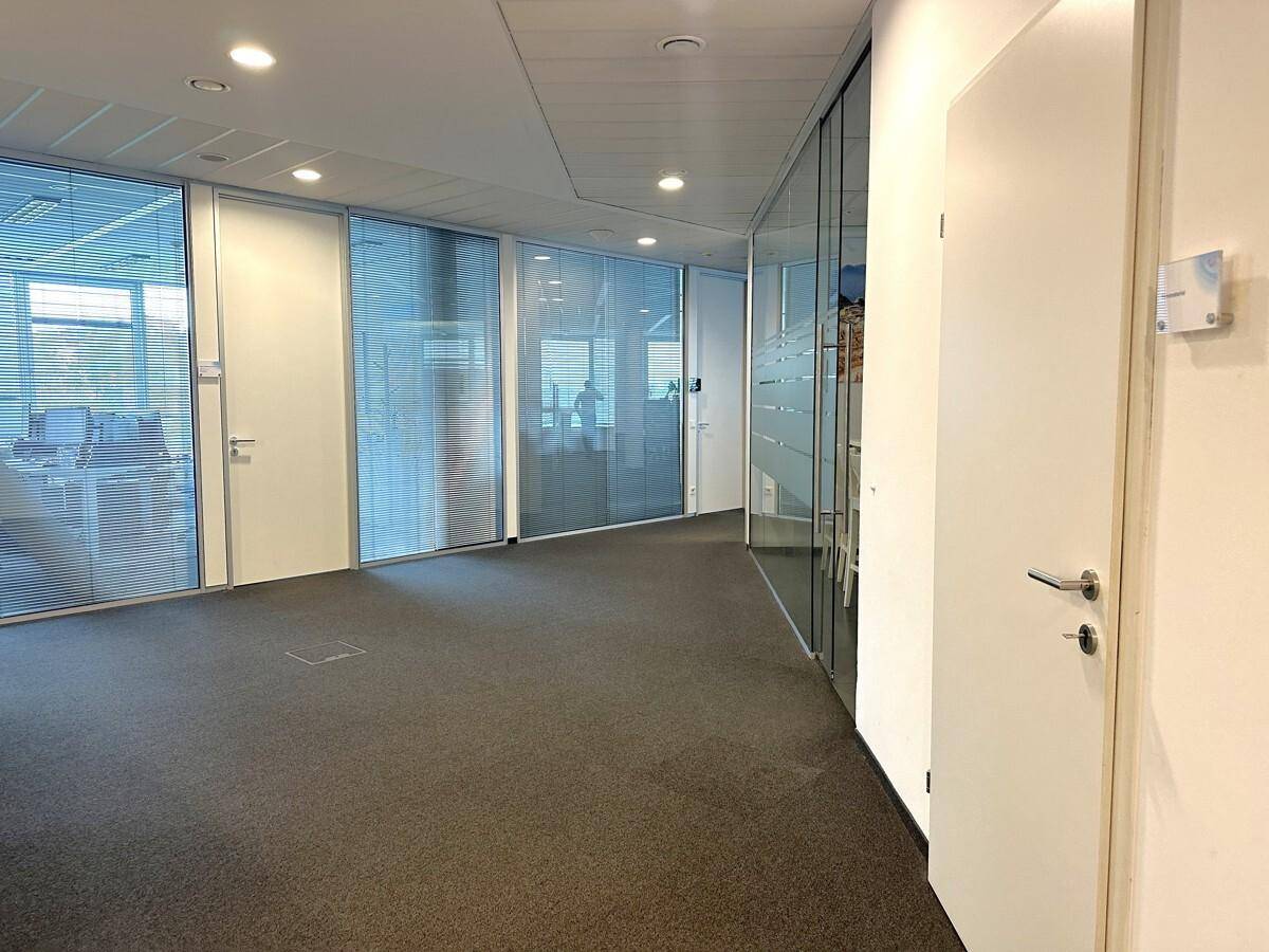 Heller Büroflur mit Teppichboden und Glaswänden, die den Blick in angrenzende Büroräume ermöglichen.