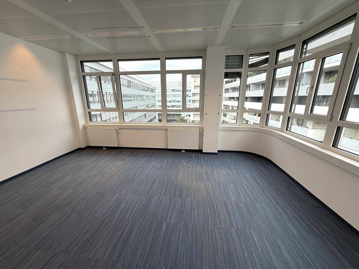 Helles Büro mit großen Fenstern, blauem Teppichboden und Blick auf umliegende Gebäude.