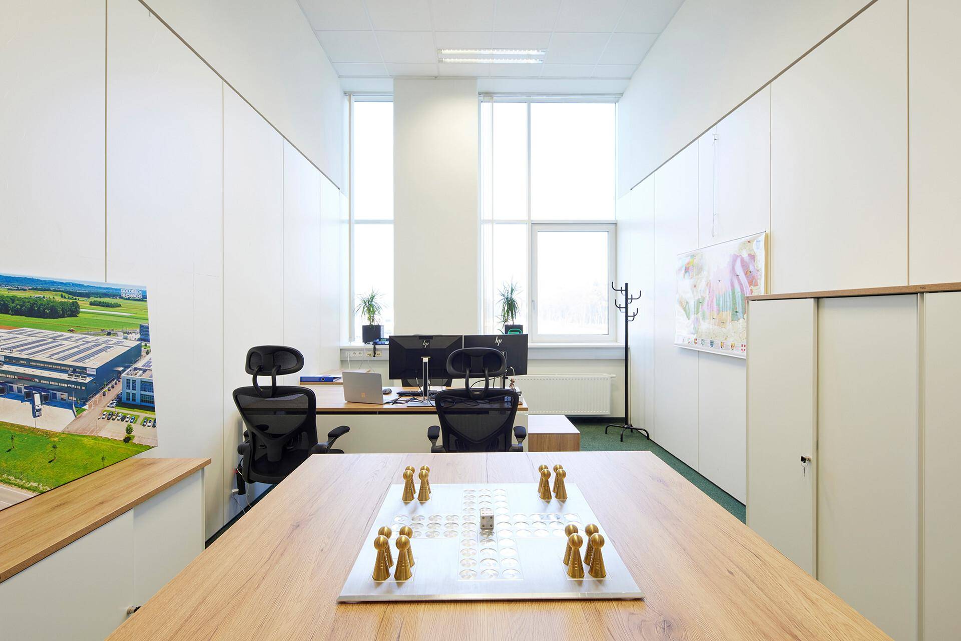 Modernes Büro mit Holztisch, zwei Arbeitsplätzen und großen Fenstern, die viel Tageslicht hereinlassen.