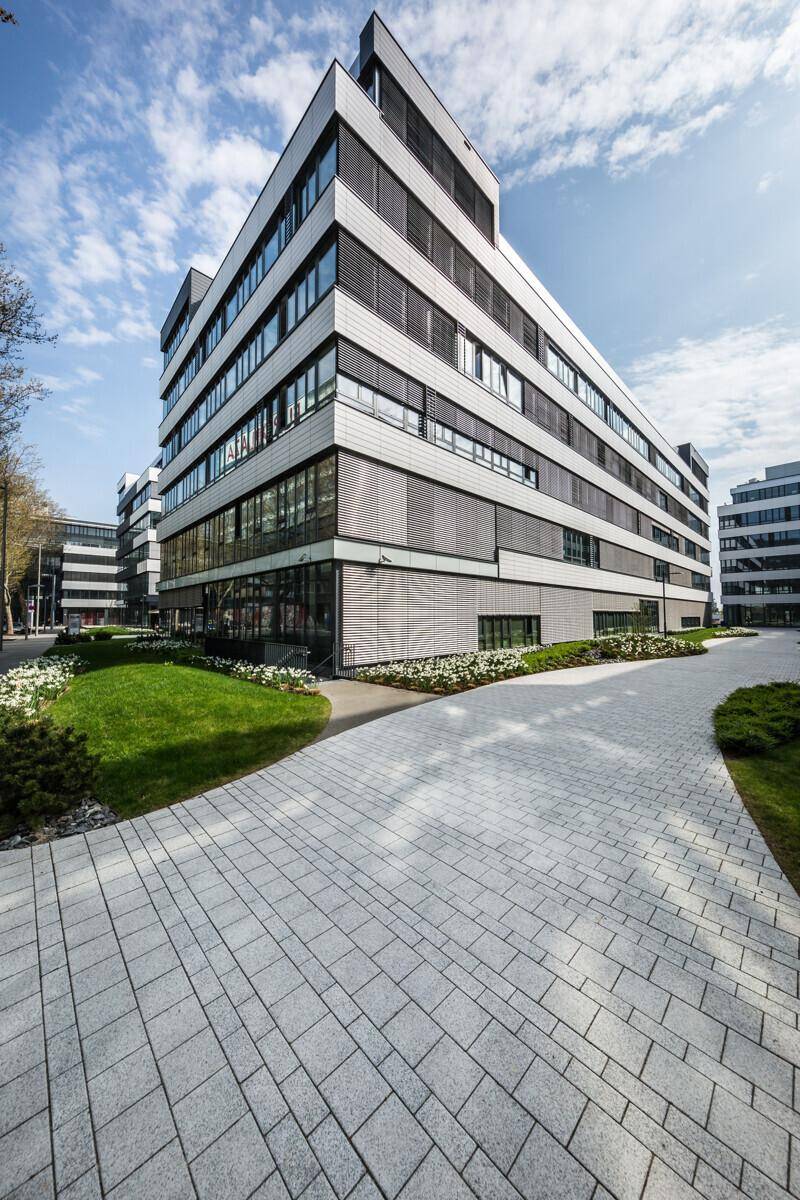 Weitläufiger gepflasterter Weg führt zu einem modernen Bürogebäude mit großen Fensterfronten.