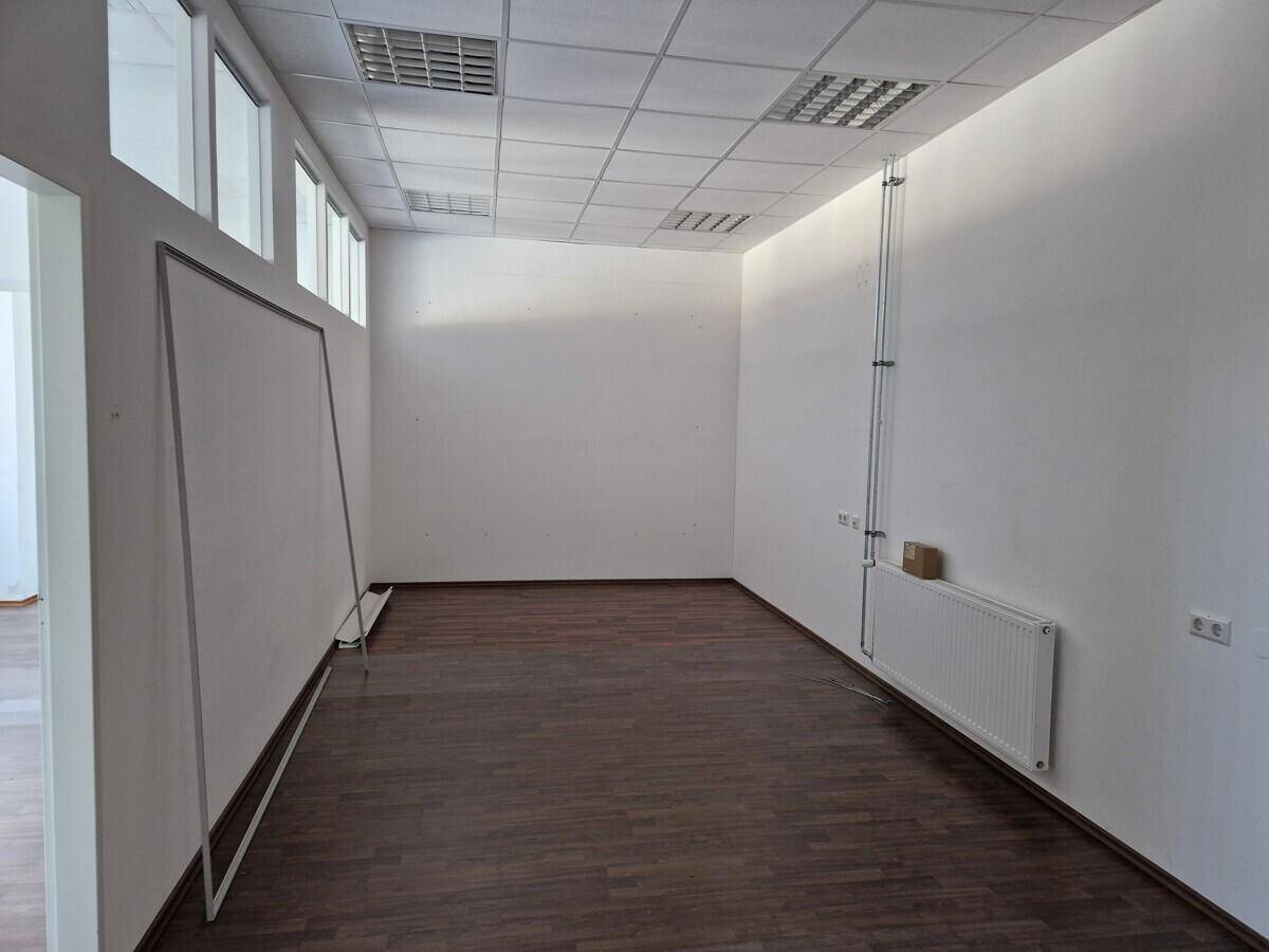 Großer, leerer Raum mit dunklem Laminatboden, weißen Wänden und einer Rasterdecke mit Beleuchtung.