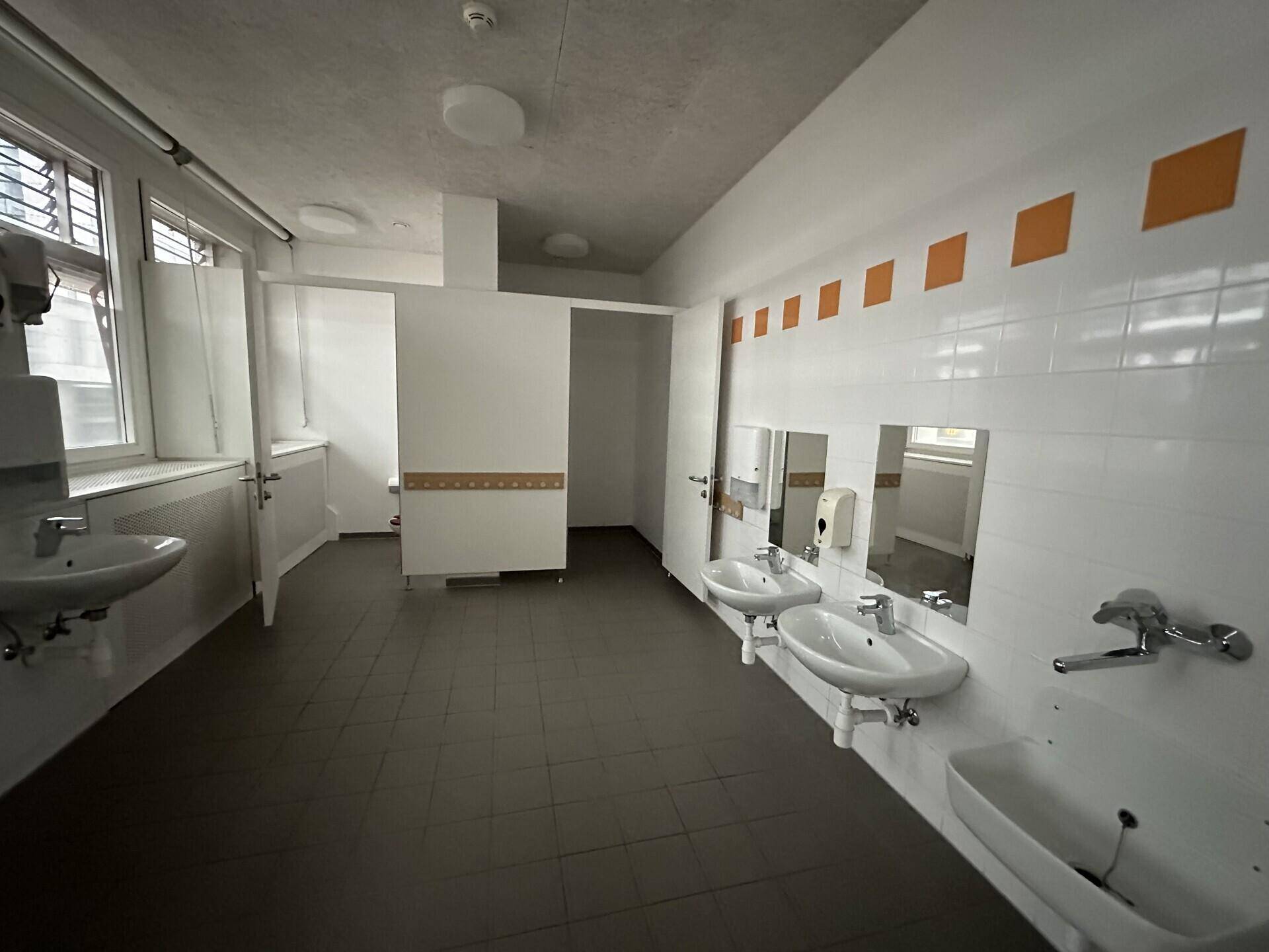 Großer Waschraum mit mehreren Waschbecken, Fliesenboden und Fenstern für natürliche Belüftung.