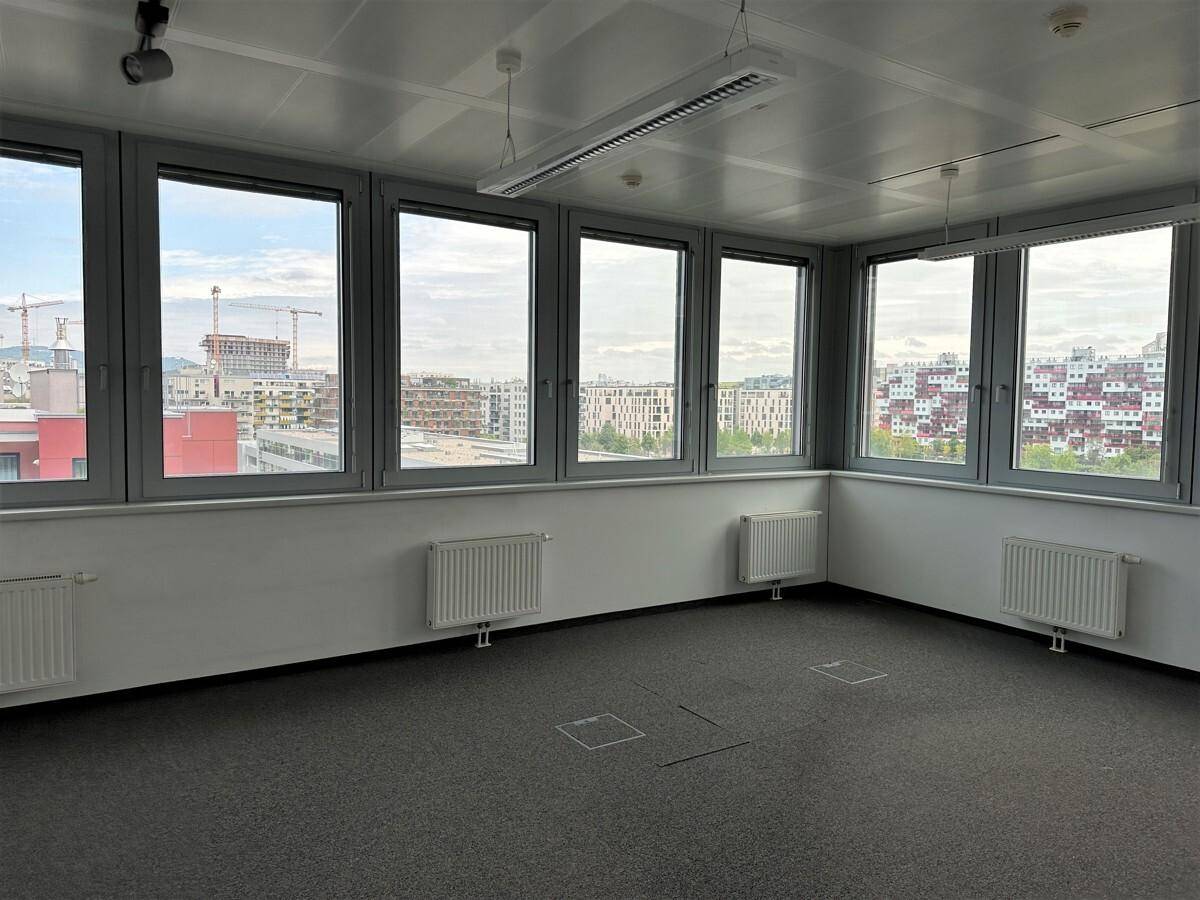 Heller Büroraum mit Teppichboden und einer Fensterfront, die einen weiten Blick über die Stadt bietet.