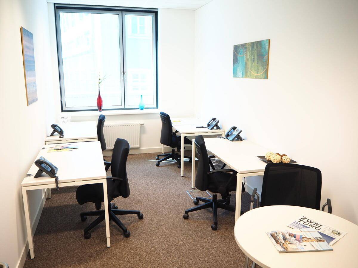 Helles Büro mit mehreren Arbeitsplätzen, großen Fenstern und Teppichboden, ideal für Coworking.