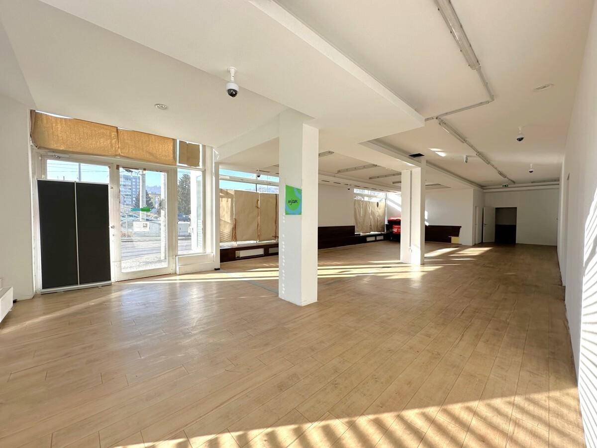 Geräumiger, leerer Ladenbereich mit großen Fenstern, Holzboden und Säulen, gut beleuchtet durch Tageslicht.