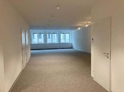 Langer Flur mit Teppichboden und mehreren Türen, der zu einem hellen Büroraum mit vielen Fenstern führt.