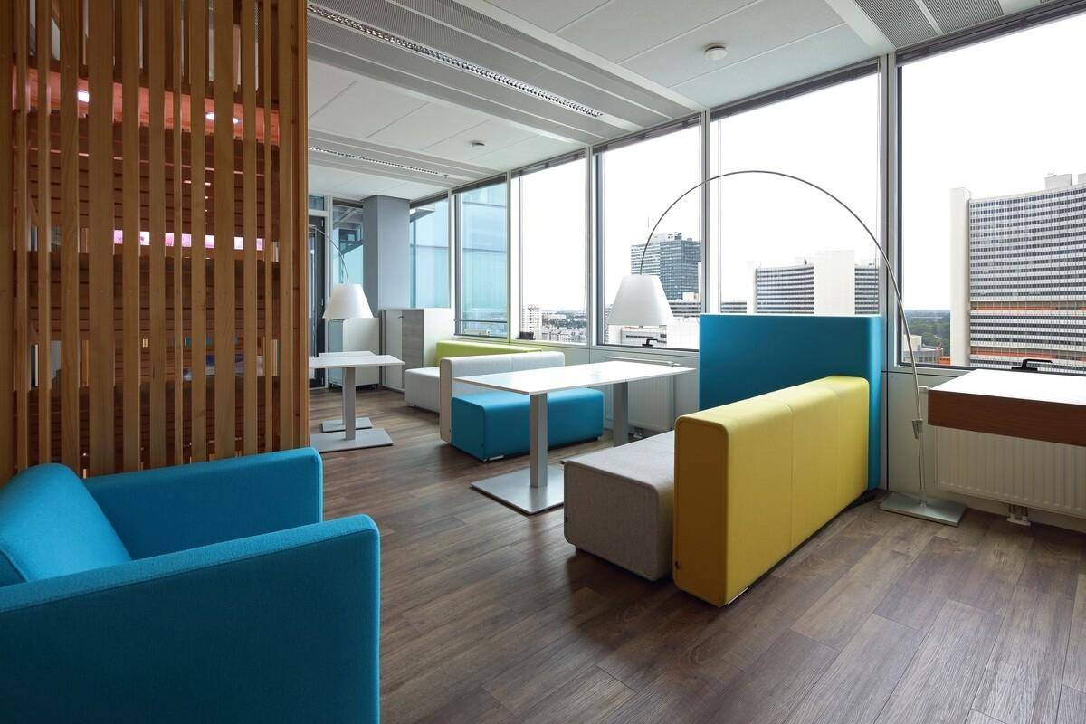 Moderne Lounge-Zone im Büro mit farbenfrohen Sitzgelegenheiten und Holztrennwänden, hell und offen.