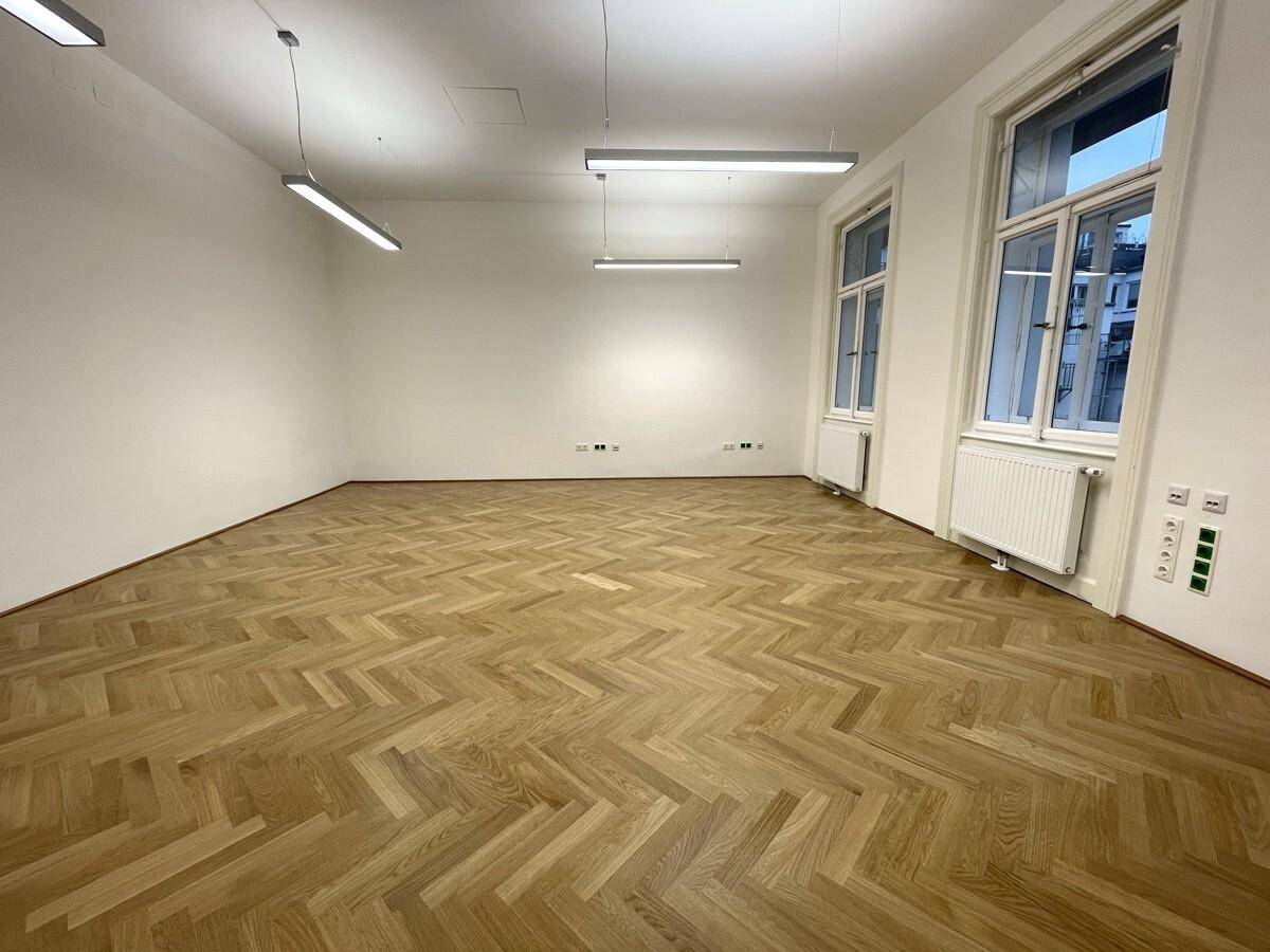 Leerer Büroraum mit klassischem Fischgrätparkett und zwei großen Fenstern, die den Raum erhellen.